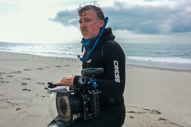 Dr. Craig O’Connell wearing dive equipment and holding an underwater camera on a beach.