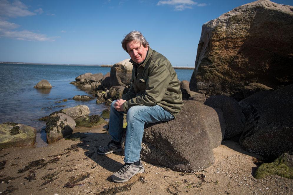 Dr. Greg Skomal seated on coastal rocks beside the ocean.
